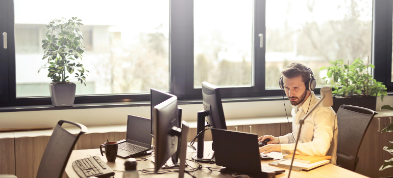 Man wearing a headset working at a desk with multiple computer monitors in a modern office setting.