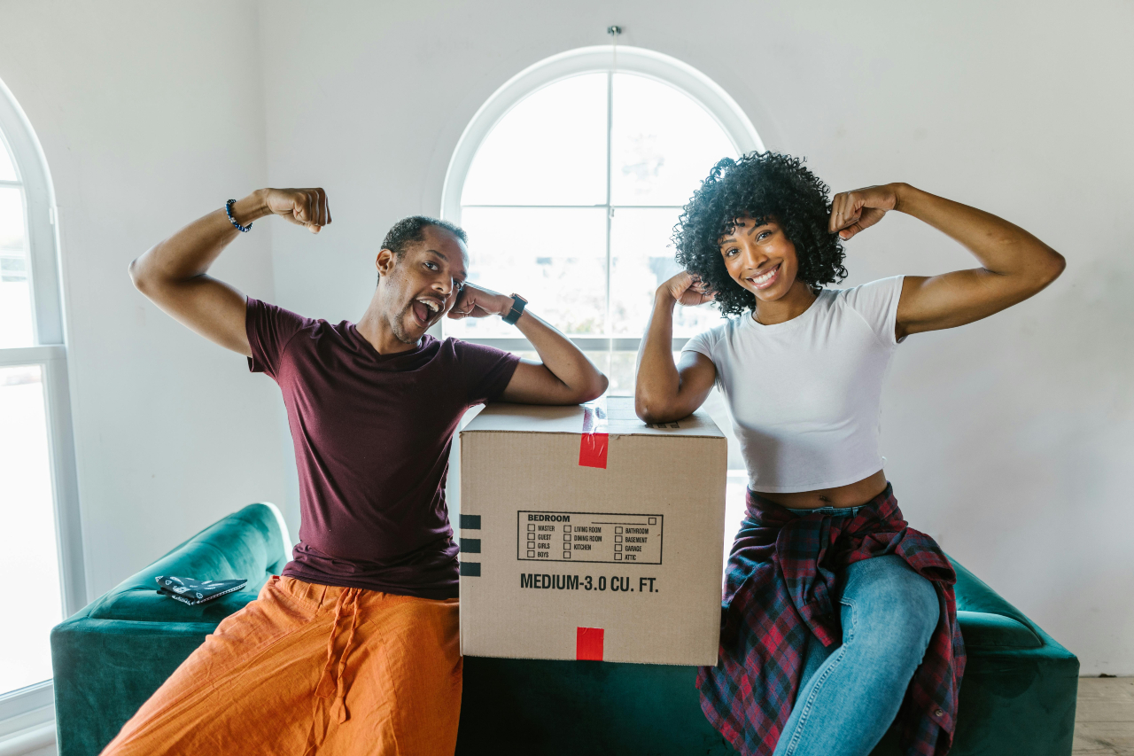 A smiling couple flexing their arms while sitting next to a medium-sized moving box in a bright room.