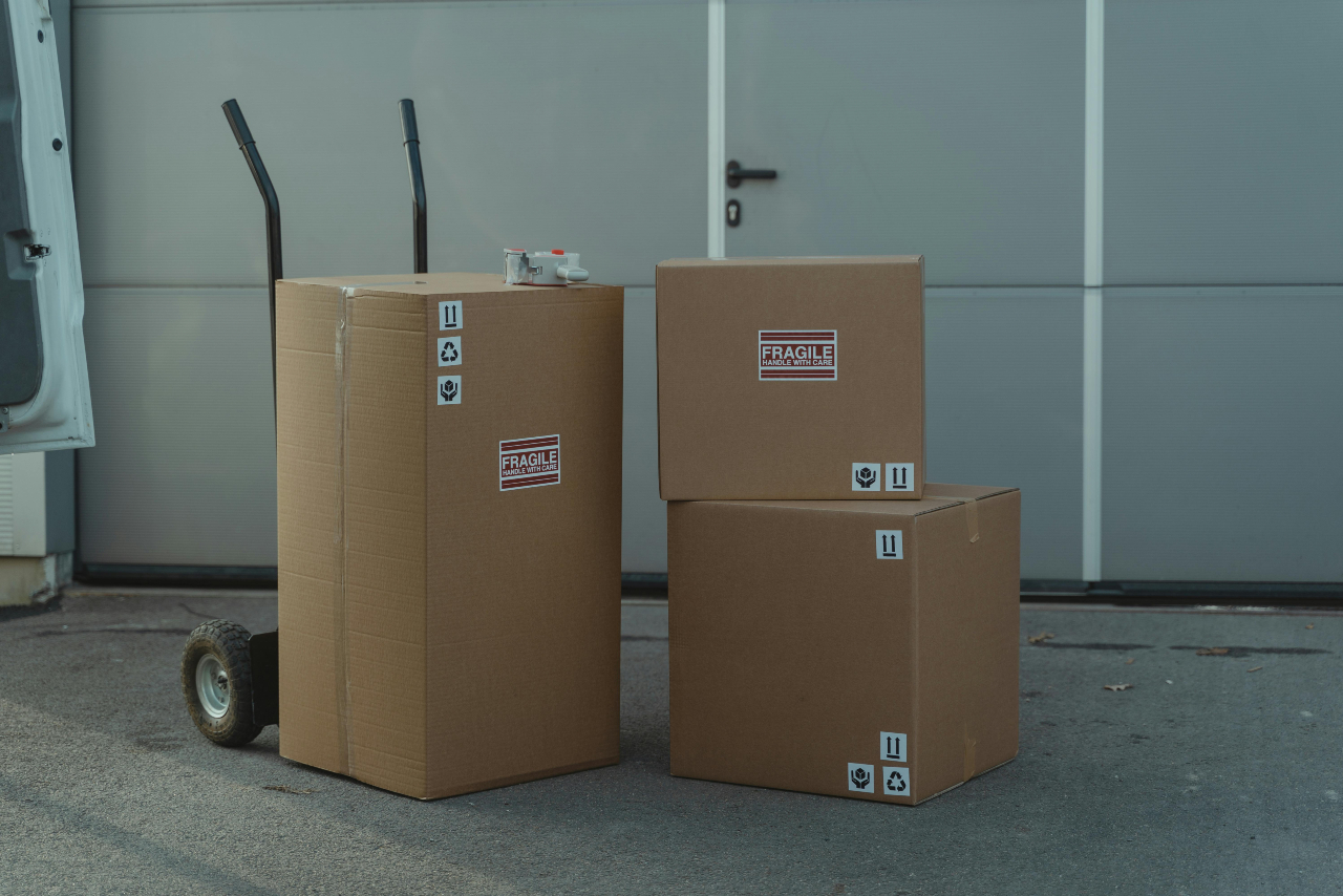 Three cardboard boxes labeled 'fragile' with eco-friendly handling symbols, placed next to a hand truck outside a garage.