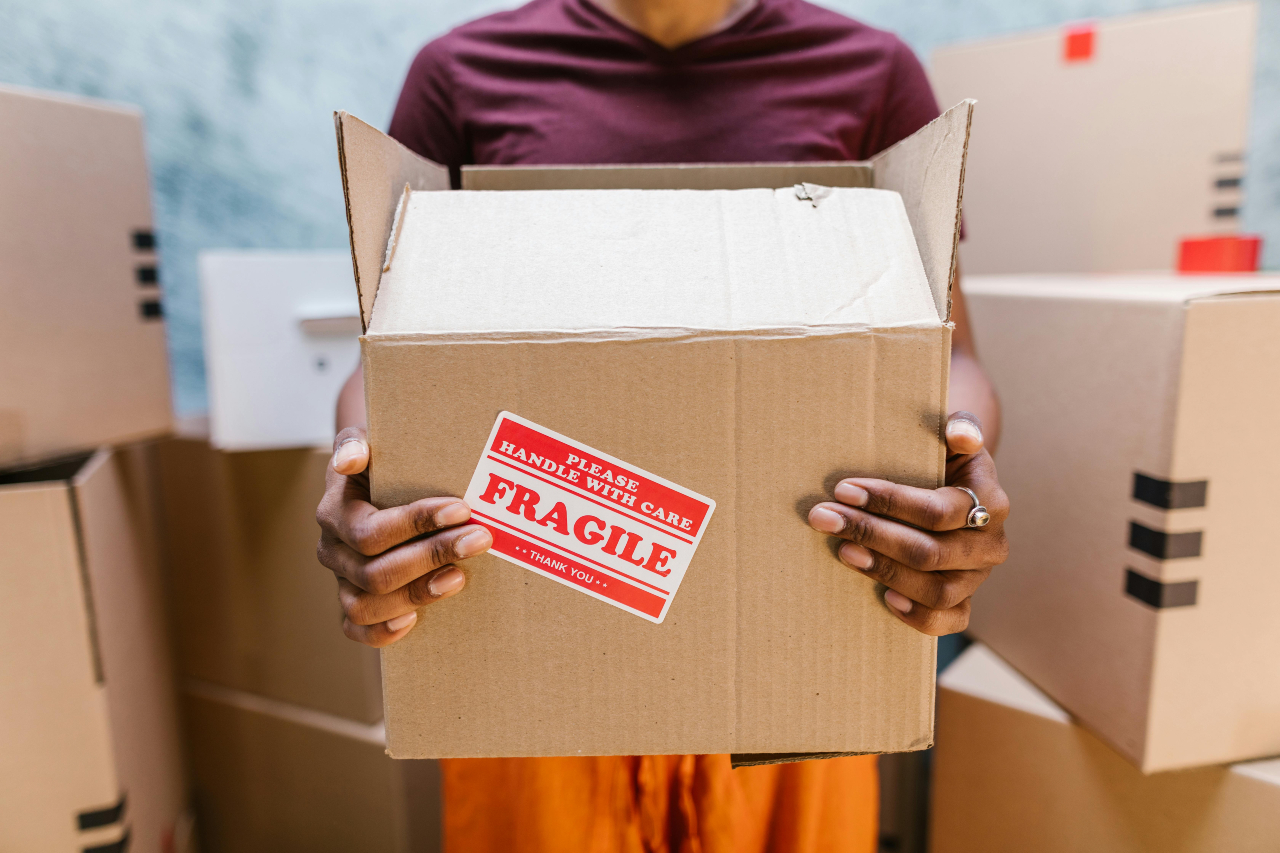A person holding a cardboard box labeled "FRAGILE" with both hands, surrounded by other packed boxes.