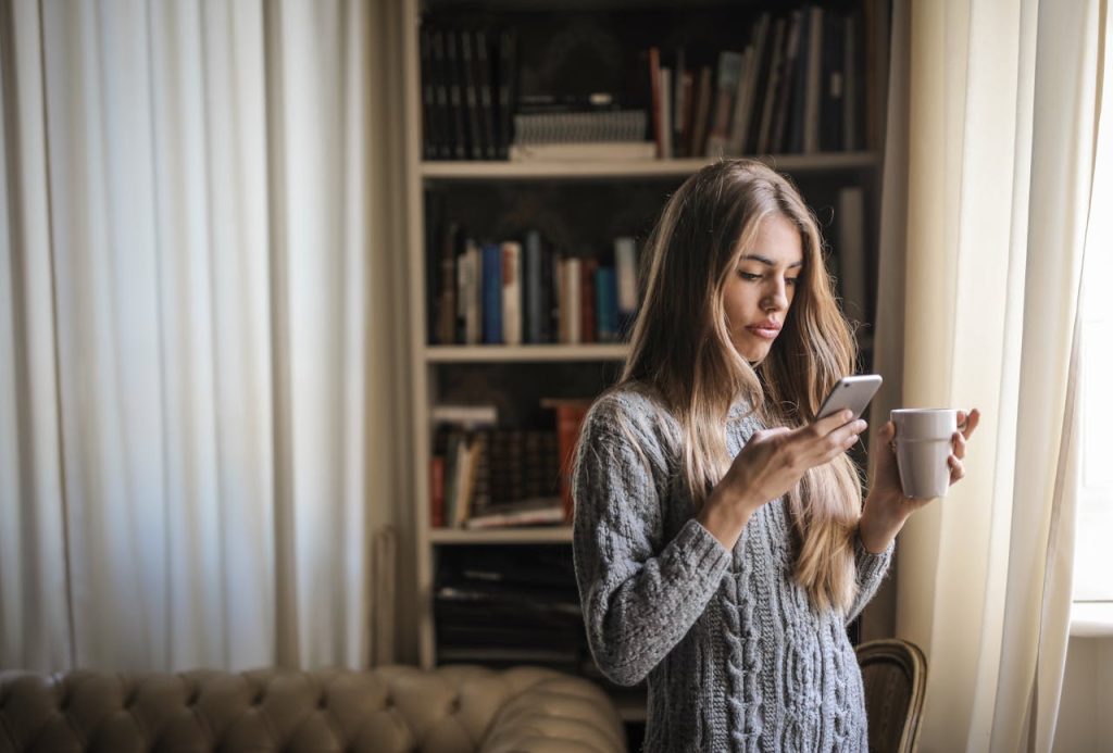 Photo of Woman in Gray Sweater Holding White Ceramic Cup While Using Her Phone and booking packing services Boston MA
