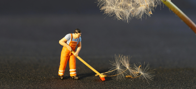 A miniature figurine in an orange uniform sweeping large dandelion seeds on a dark surface.