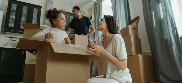 A family unpacking boxes together in a bright room, with a child smiling and drawing on a cardboard box.
